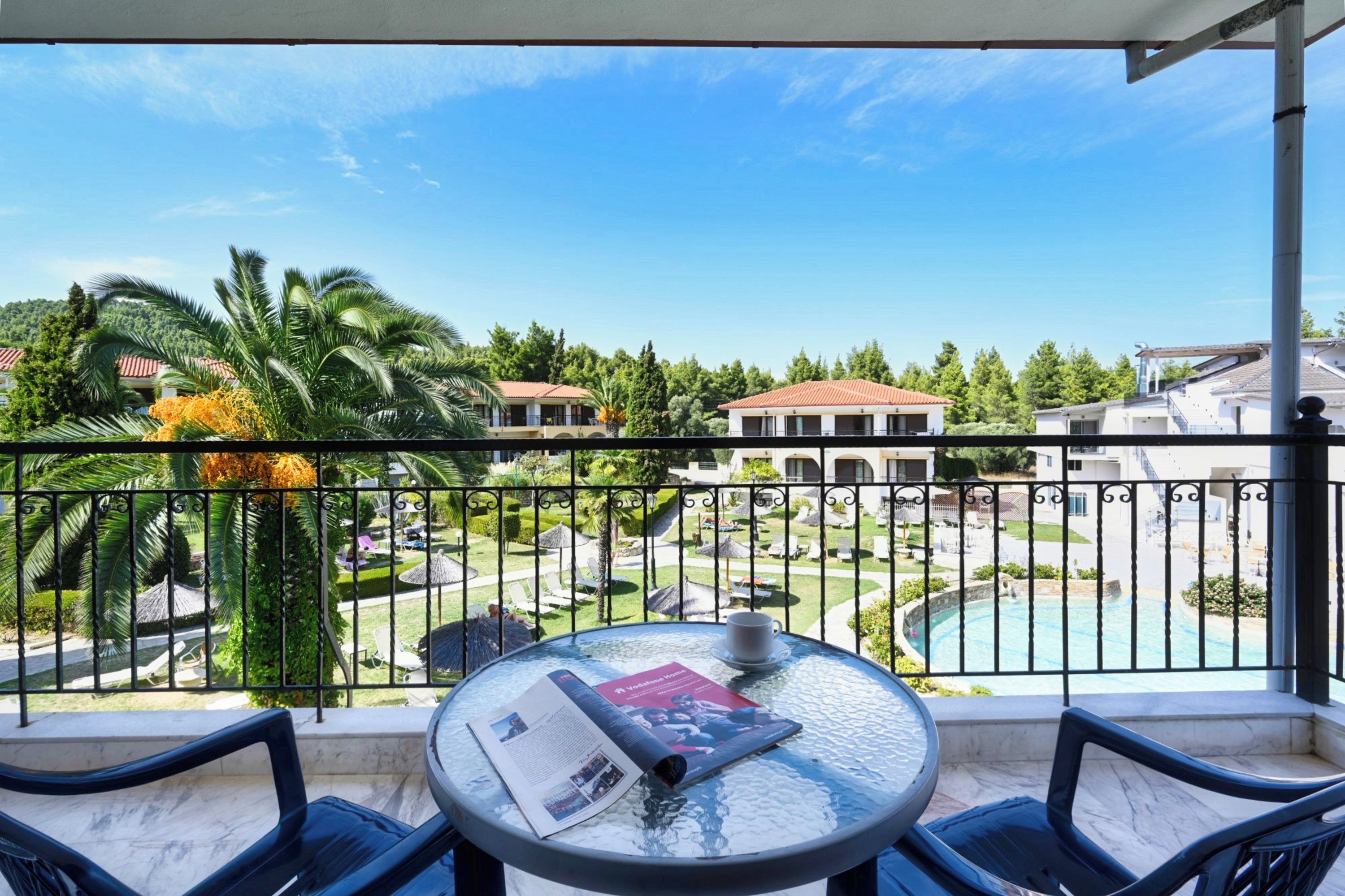 A balcony with a glass table, two chairs, and a view of a pool and resort buildings.