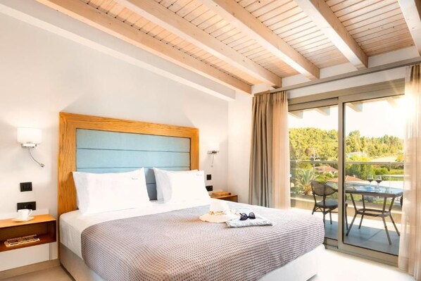 A hotel room with a large bed, a wooden headboard, a balcony with a table and chairs, and a view of greenery. - Chrousso Village (Kassandra)