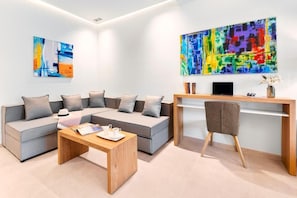 A modern living room with a grey sectional sofa, a wooden coffee table, and a desk with a chair. There are abstract paintings on the walls and a laptop on the desk. - Chrousso Village (Kassandra)
