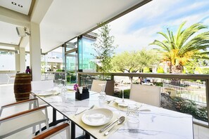 Un coin repas avec une table en verre dressée pour deux personnes, donnant sur un jardin planté de palmiers et offrant une vue sur le paysage urbain.