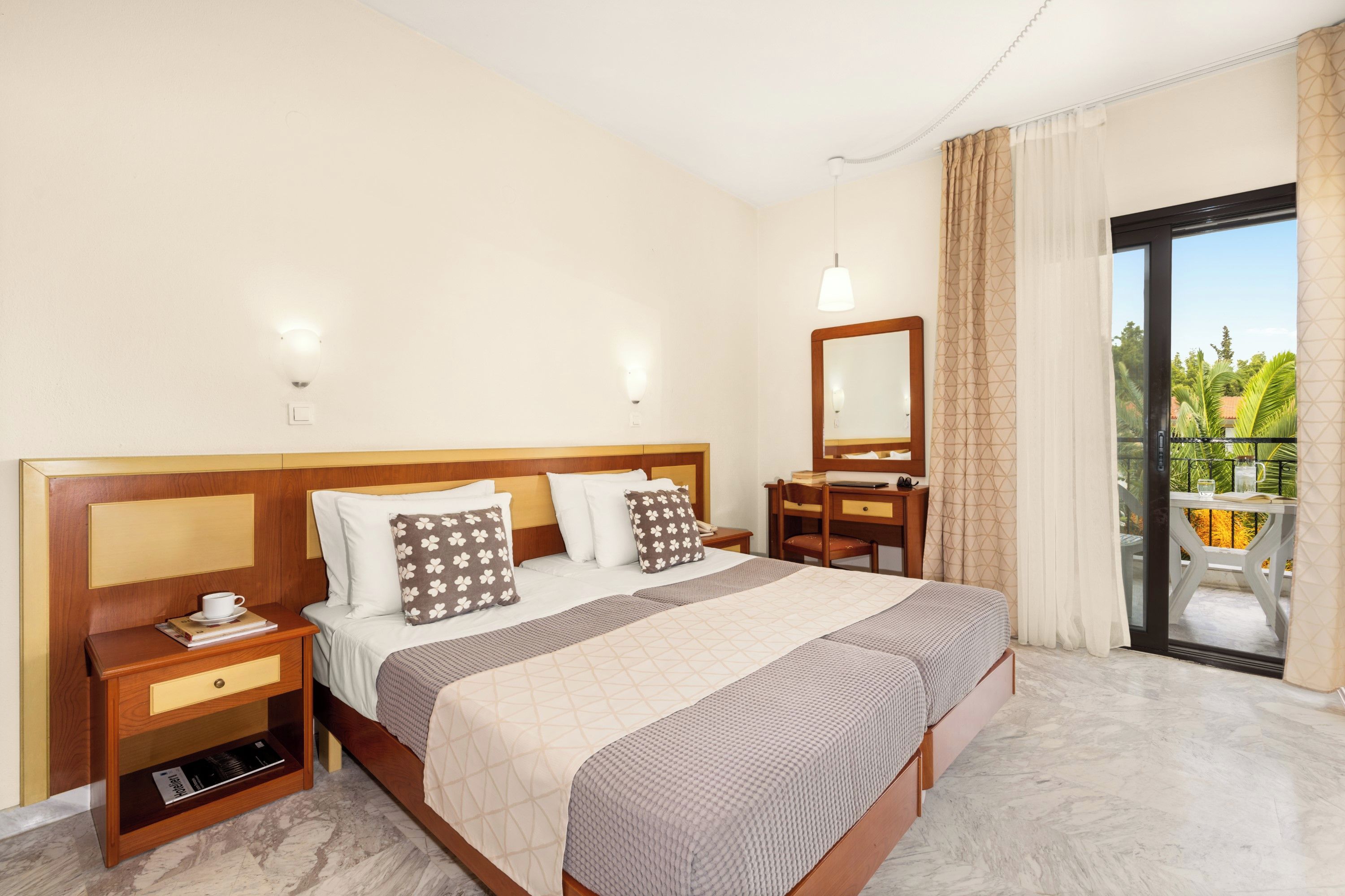 A hotel room with a large bed, a desk, a chair, a small table, and a balcony with a view of greenery.