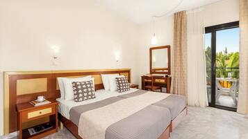 A hotel room with a large bed, a desk, a chair, a small table, and a balcony with a view of greenery.
