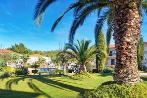 A well-maintained outdoor area with palm trees, a grassy lawn, and a building with a red roof. - Chrousso Village (Kassandra)