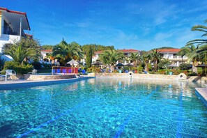 A clear blue pool surrounded by palm trees and buildings with red-tiled roofs. - Chrousso Village (Kassandra)