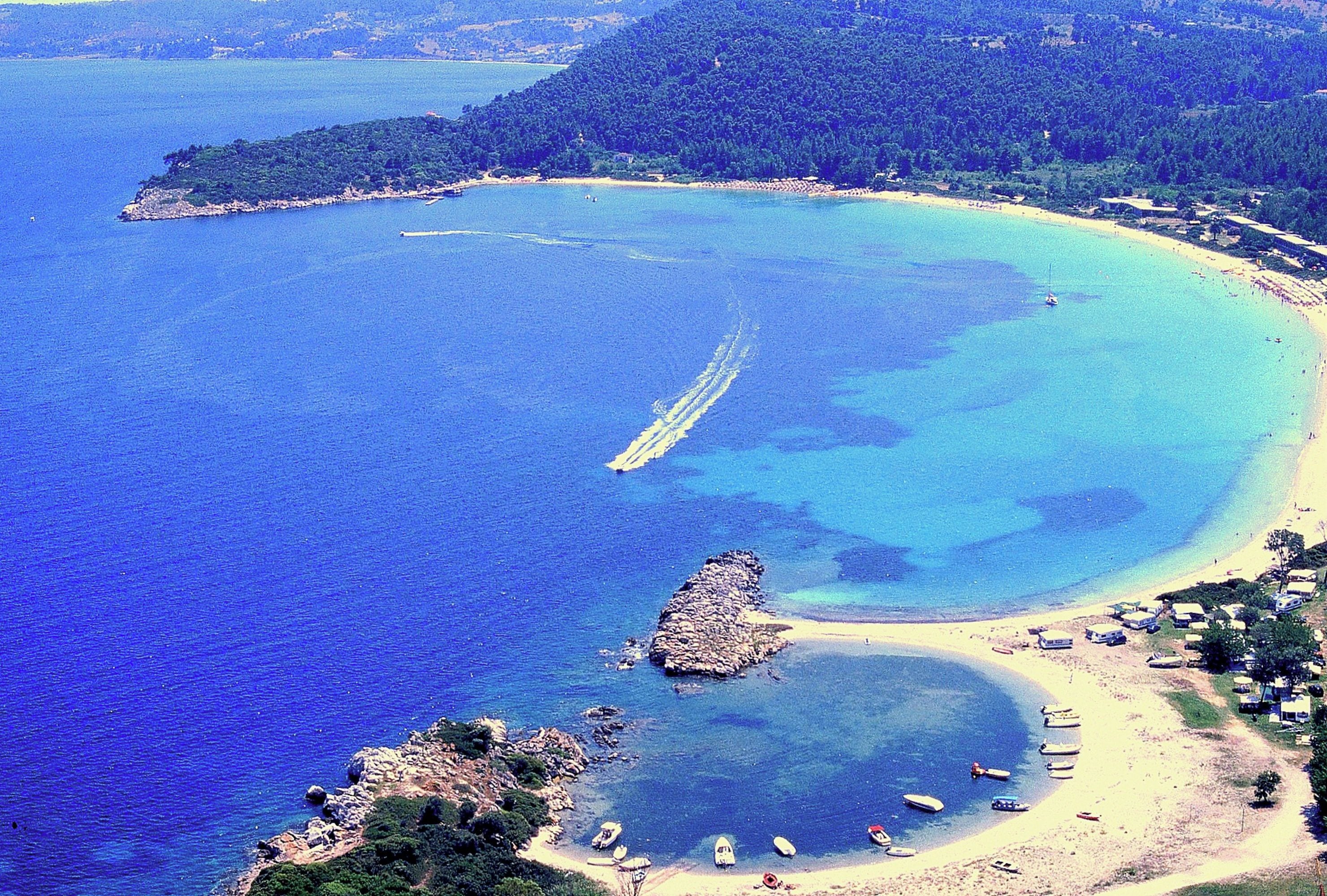 Luchtbeeld van een kustgebied met helderblauw water, een zandstrand en een klein rotsachtig eiland.