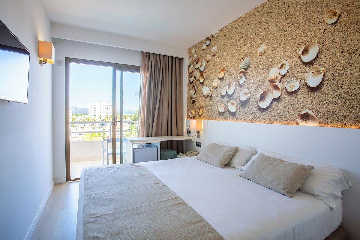 A hotel room with a large bed, a desk with a chair, and a view of the city through the balcony doors.