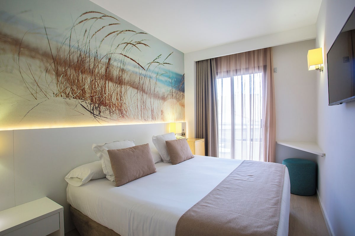 A modern hotel room with a large bed, a painting of tall grasses, a flat-screen TV, and a window with curtains.