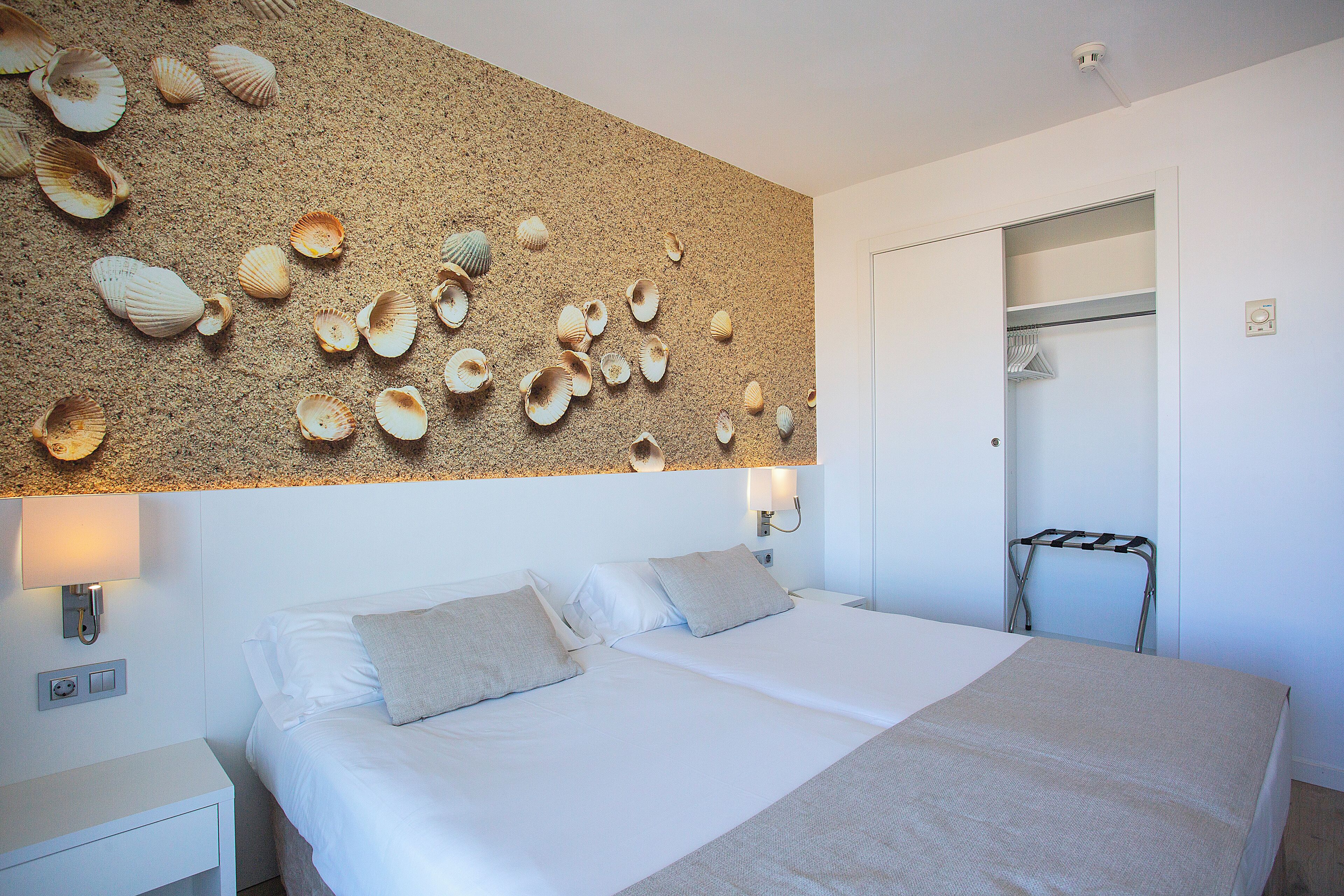 Un dormitorio con una cama, dos mesitas de noche, un estante en la pared con conchas decorativas, una lámpara de pared y un armario con un tendedero.