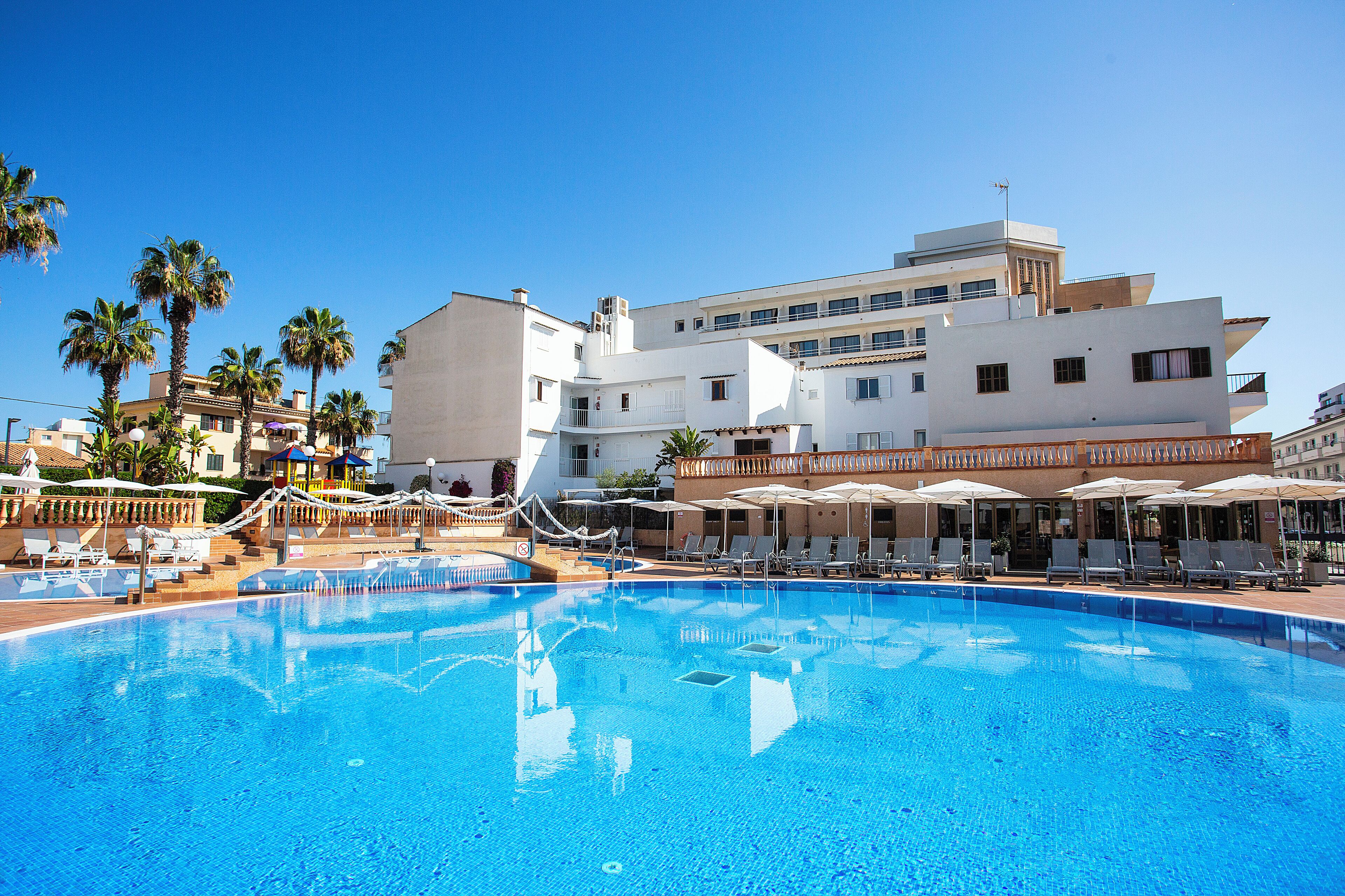 Una piscina con sillas de descanso y sombrillas, rodeada de edificios con balcones y palmeras.