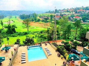 Zona de piscina con sillones reclinables y sombrillas, rodeada de follaje y un campo de golf al fondo.