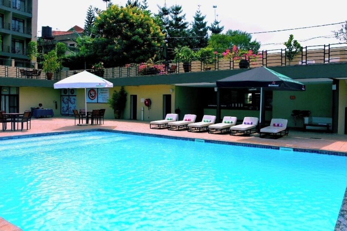 Una piscina con sillas de descanso y sombrillas, rodeada por un edificio con balcones y plantas en macetas.