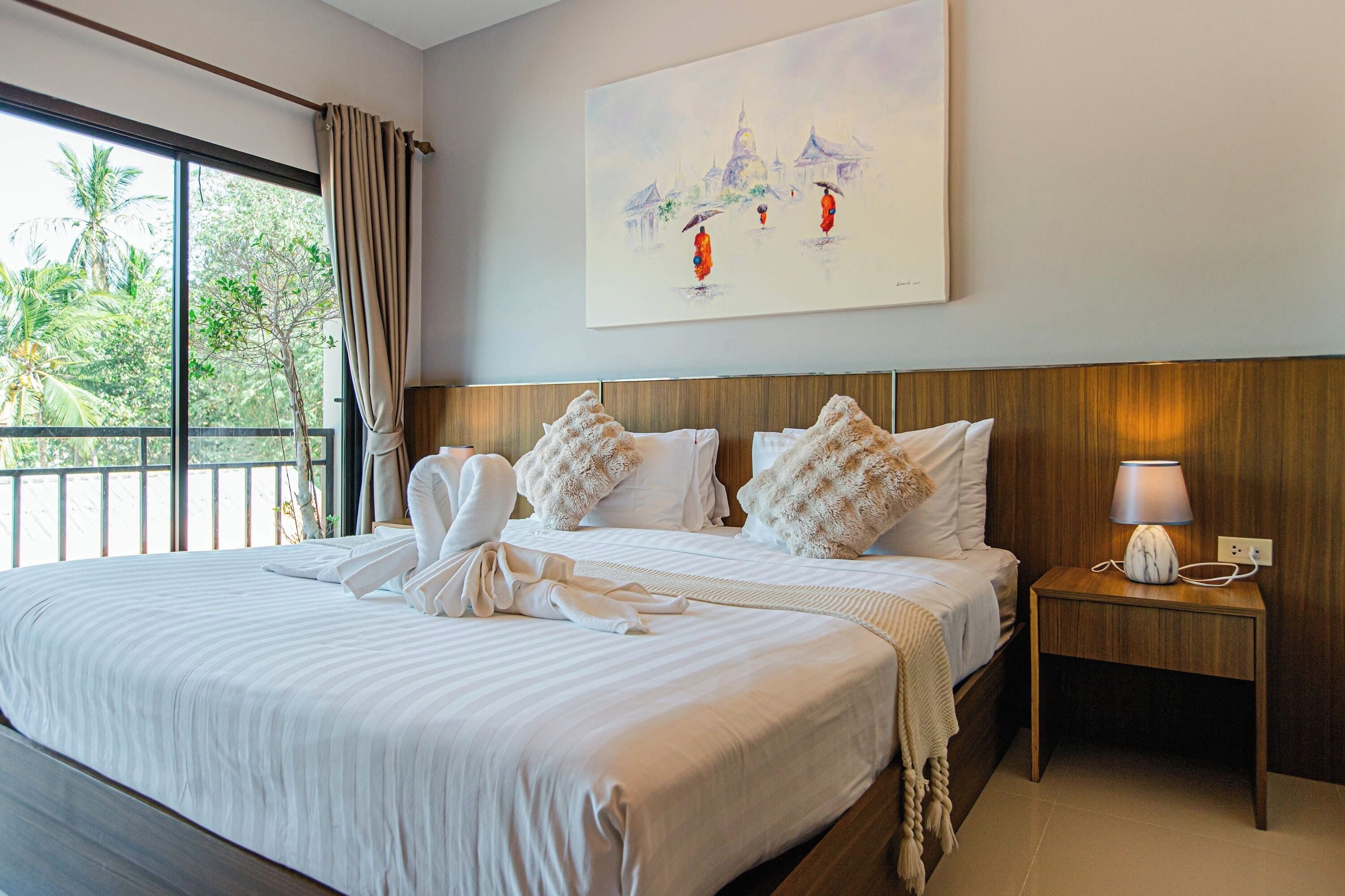 Habitación de hotel con una cama grande, una mesita de noche con lámpara, un cuadro en la pared y vistas a árboles por la ventana.