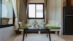 A room with a window, a sofa with cushions, a table with items, and a refrigerator. - Happy Eight Resort (Rawai)