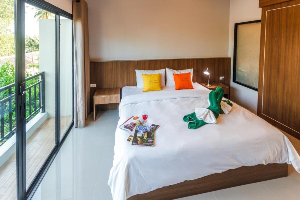 A hotel room with a large bed, a nightstand, a lamp, and a view of greenery outside.