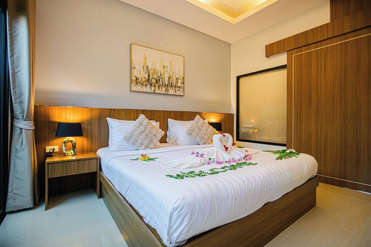 A hotel room with a large bed, a nightstand with a lamp, a cityscape painting on the wall, and a wooden wardrobe.