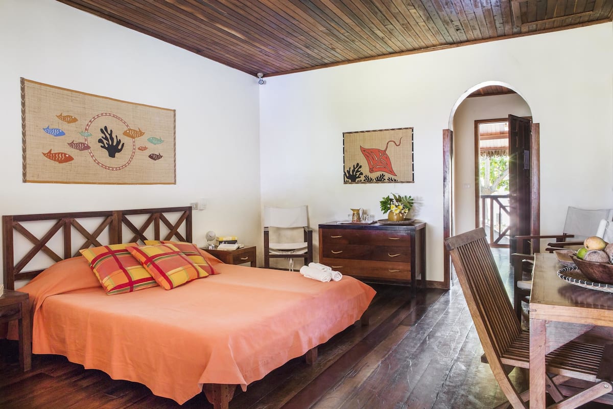 A room with a bed, wooden headboard, a wooden dresser, a chair, and a table with a bowl of fruit.