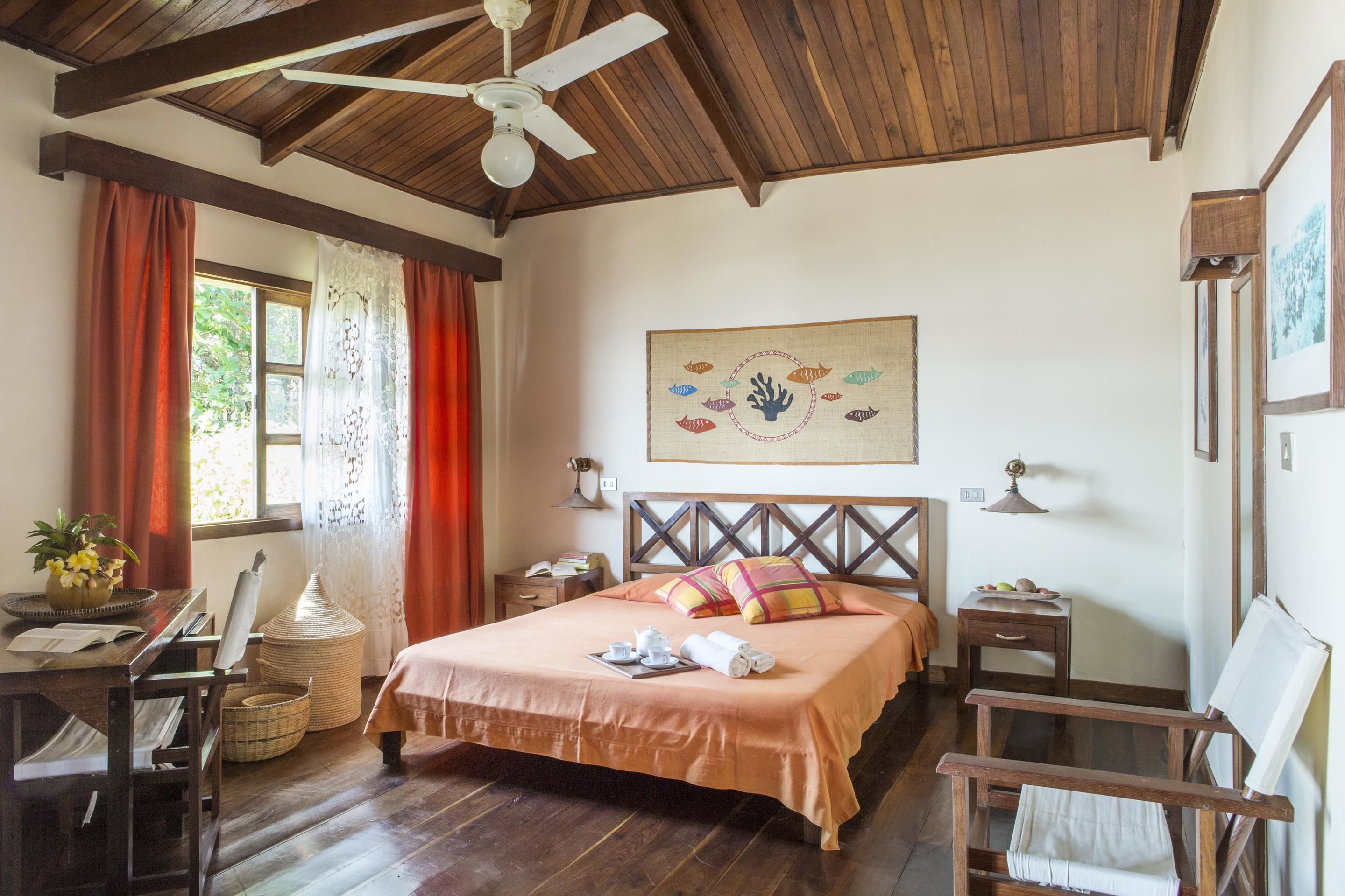 A well-lit bedroom with a wooden ceiling, a bed with a headboard, a desk with a chair, a window with curtains, and a painting on the wall.