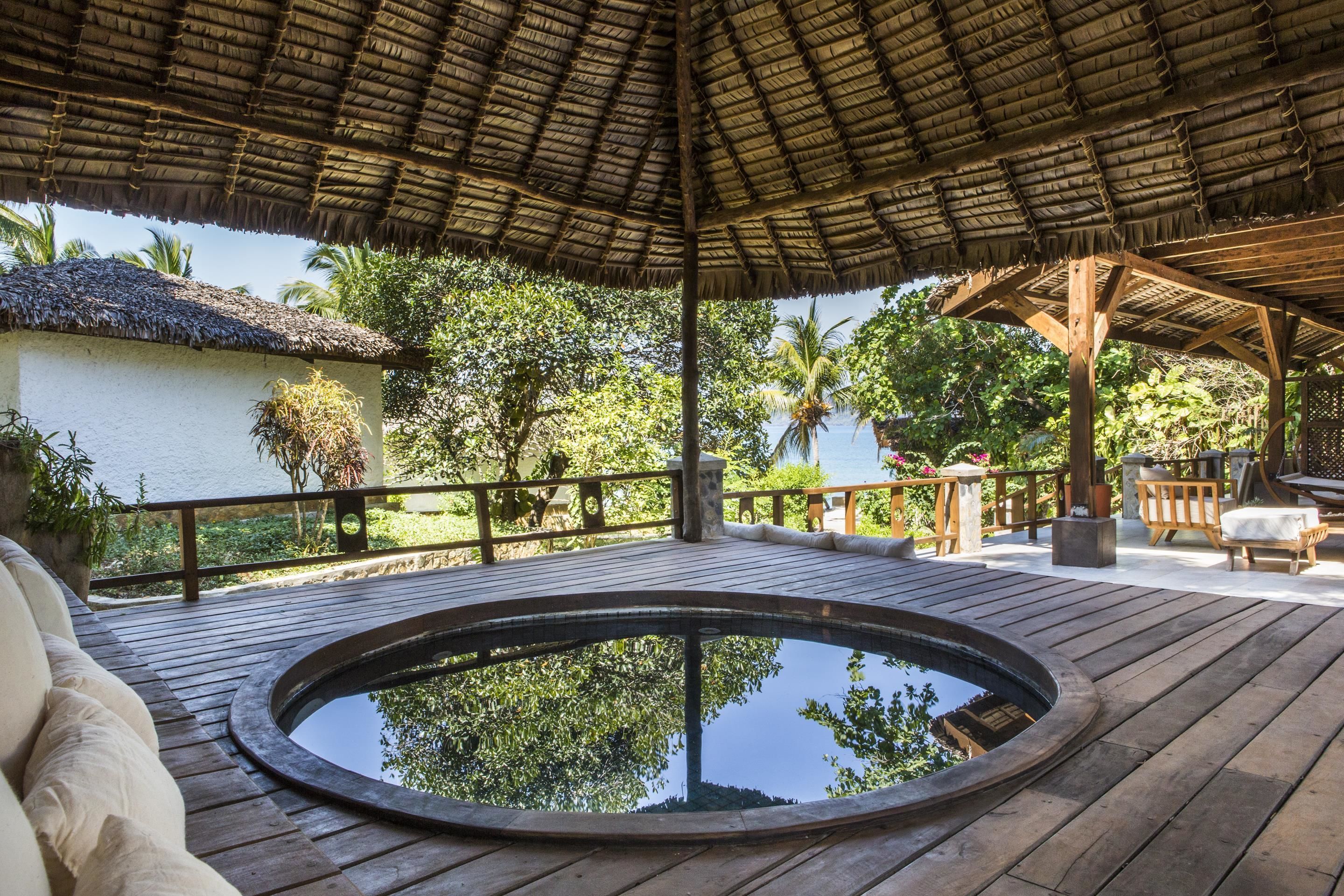 A wooden deck with a circular hot tub, a thatched roof, and a view of a white building and palm trees.