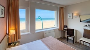 Quarto de hotel com cama, escrivaninha, cadeira, televisão e janela com vista para a praia.