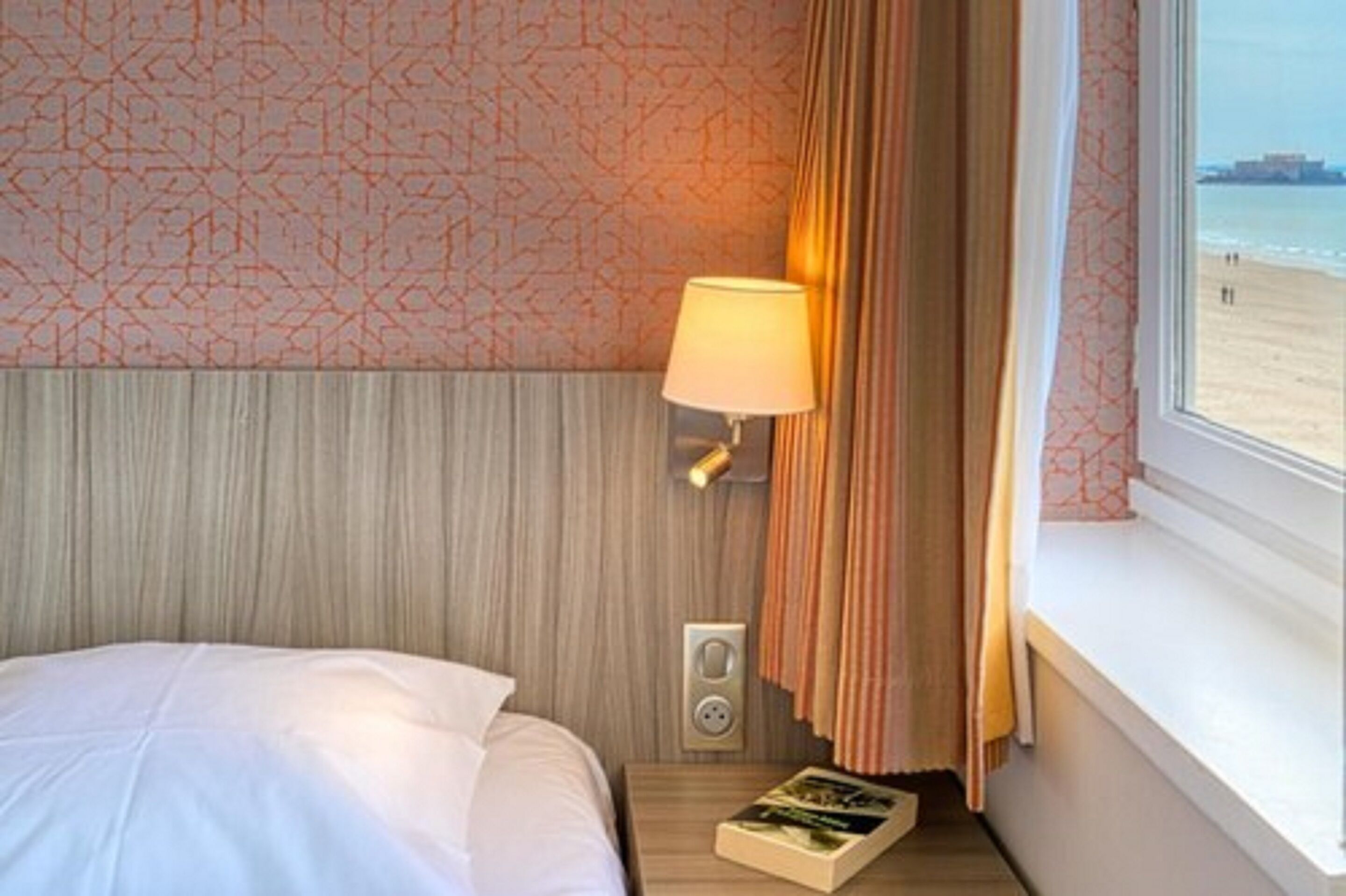 Et hotelværelse med en seng, et natbord med lampe, et vindue med gardiner og udsigt til stranden.