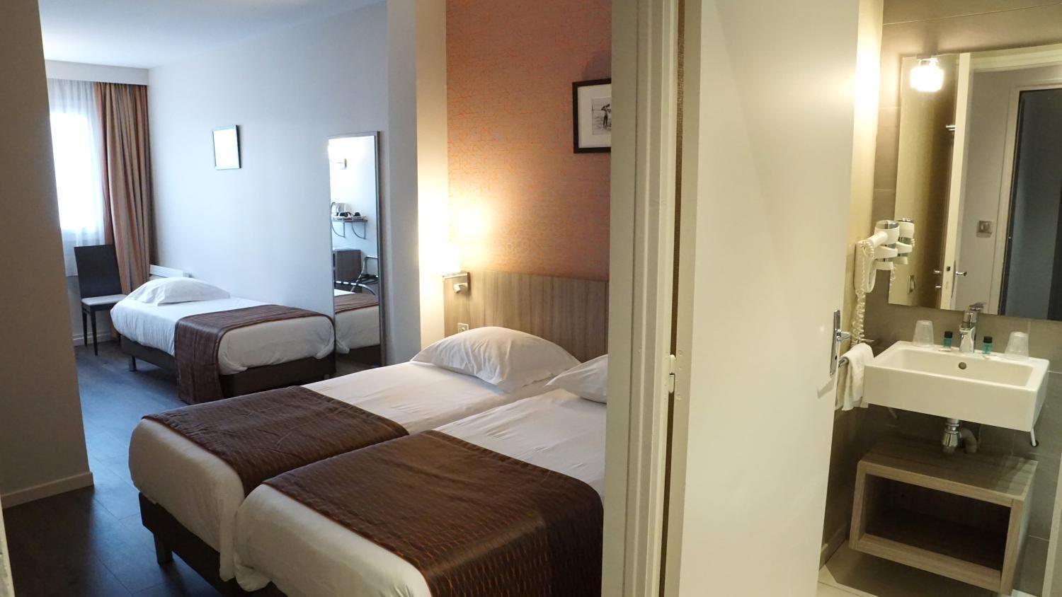 A hotel room with two beds, a desk, a chair, a bathroom with a sink and mirror.