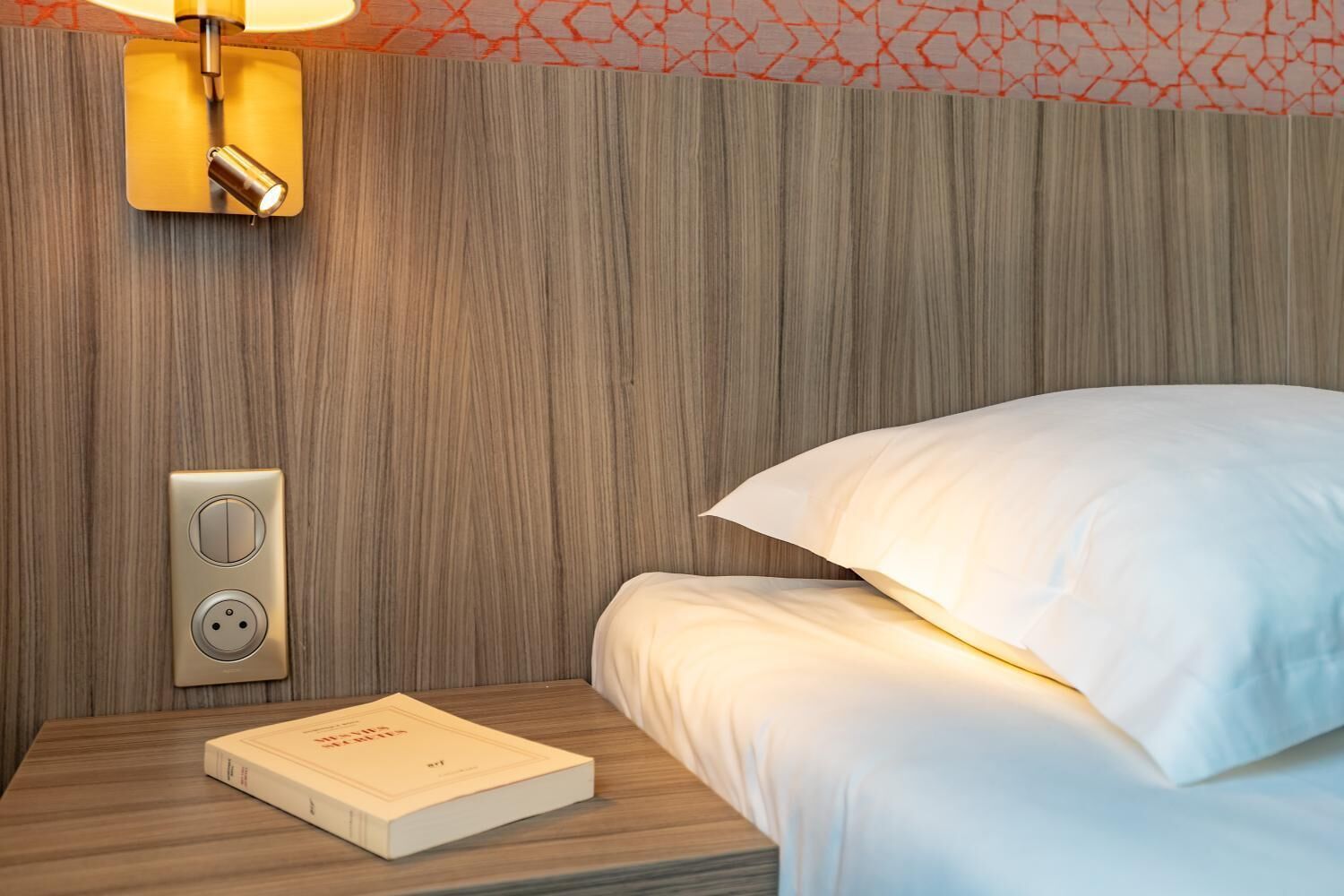 A neatly made bed with white linens, a bedside table with a book, a wall-mounted light fixture, and a power outlet.