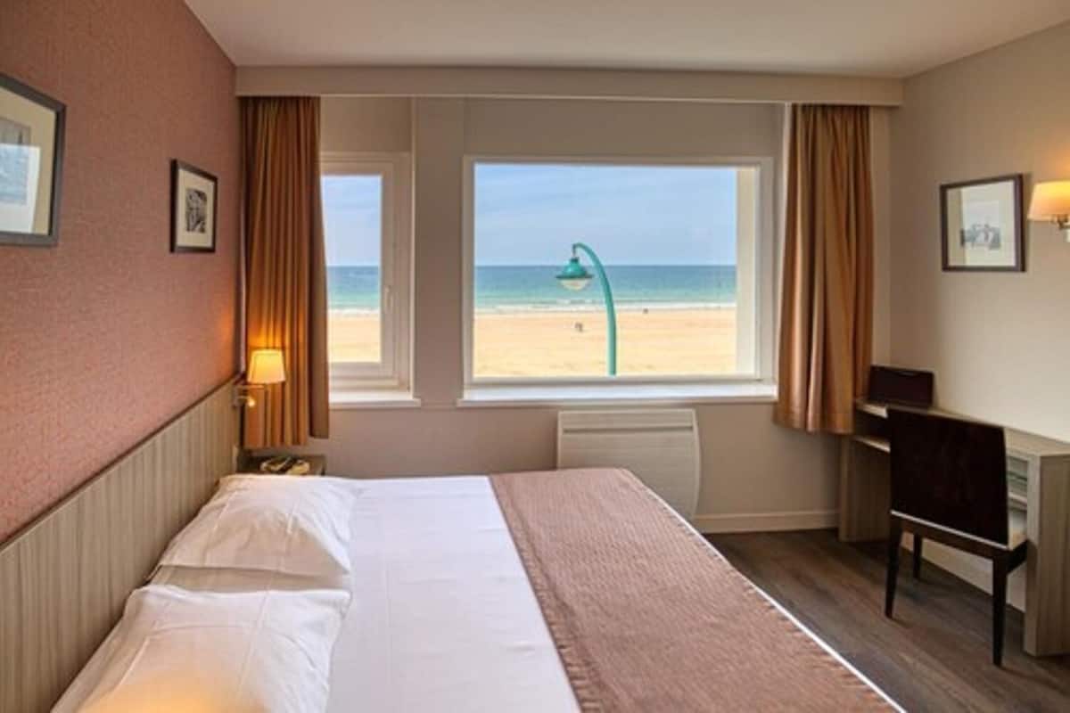 A hotel room with a bed, a desk, a chair, and a view of the beach.
