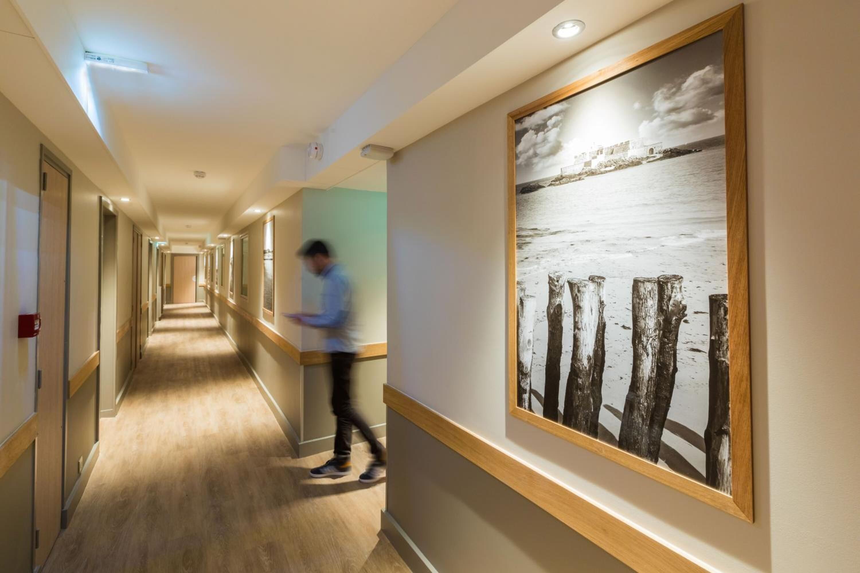 A hotel corridor with wooden flooring, a large framed artwork of trees and water on the right wall, and a person walking down the hallway.