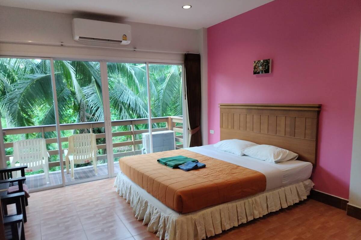 A hotel room with a bed, a balcony with chairs, and a view of palm trees.