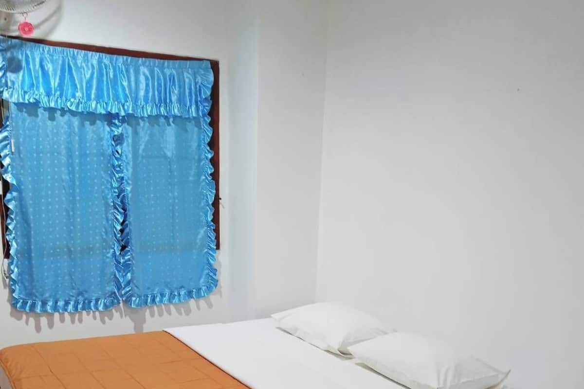 A room with a bed, a window with blue curtains, a fan, and a wooden floor.