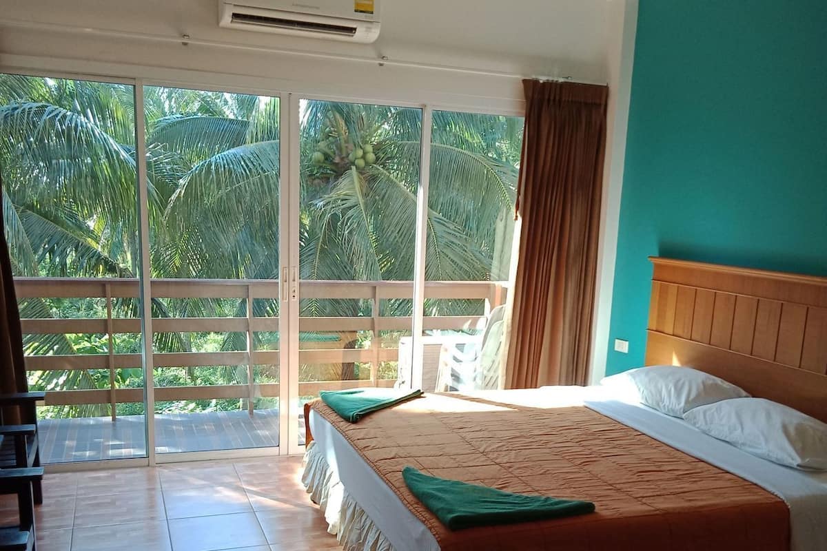 A hotel room with a bed, a balcony with a view of palm trees, and an air conditioning unit.