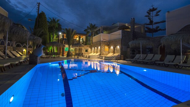 A swimming pool with lounge chairs and thatched umbrellas, surrounded by stone walls and palm trees. - Polydefkis Apartments (Santorini)