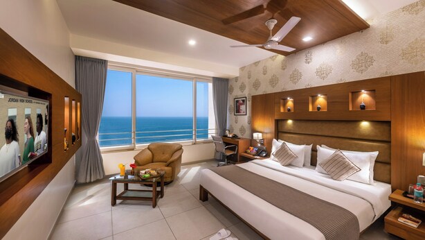 Ein Hotelzimmer mit einem großen Bett, einem Schreibtisch, einem Sessel, einem Fernseher und Blick aufs Meer.