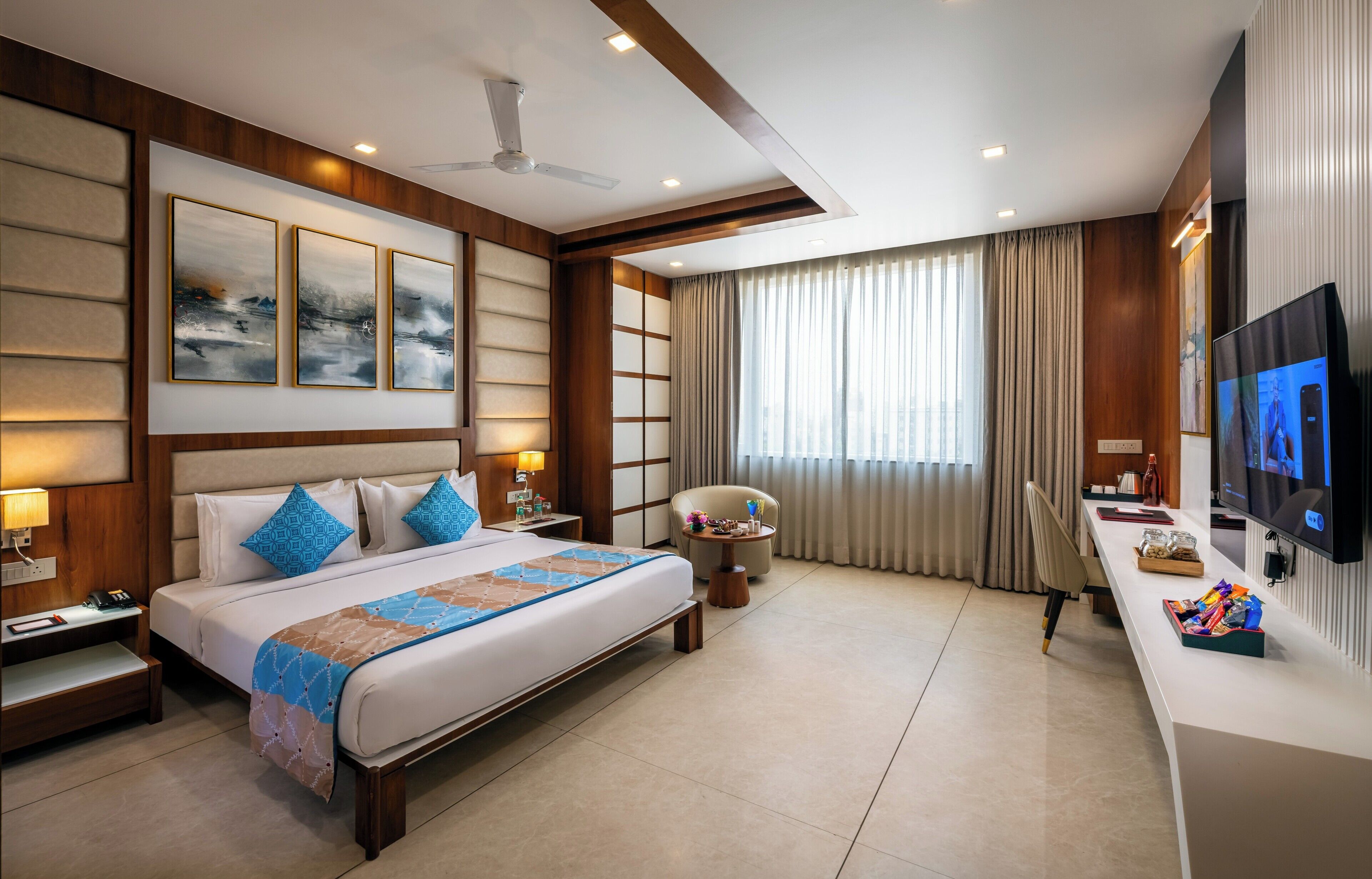 A hotel room with a large bed, bedside tables, a desk, a chair, a TV, and a ceiling fan.