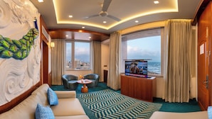 Ein modernes Zimmer mit gewölbter Decke, Deckenventilator, großem Fenster mit Vorhängen, Fernseher, zwei Sessel, eine Couch und einem gemusterten Teppich.