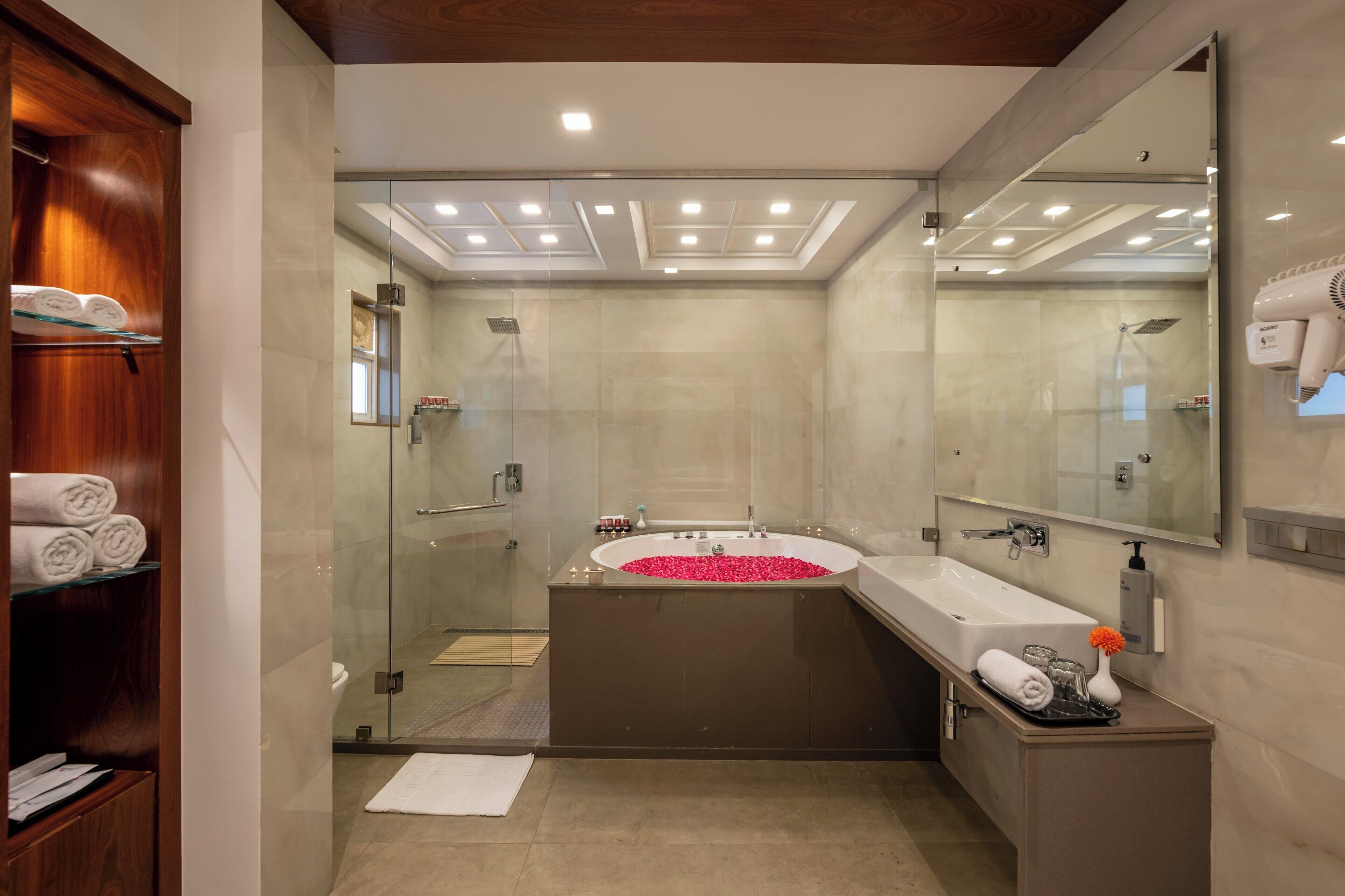 A modern bathroom with a glass-enclosed shower, a freestanding bathtub filled with rose petals, a large mirror, and a towel rack.