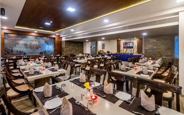 Un restaurant avec plusieurs tables dressées pour accueillir les clients, une grande fresque murale et un écran de télévision diffusant une chaîne d’information.