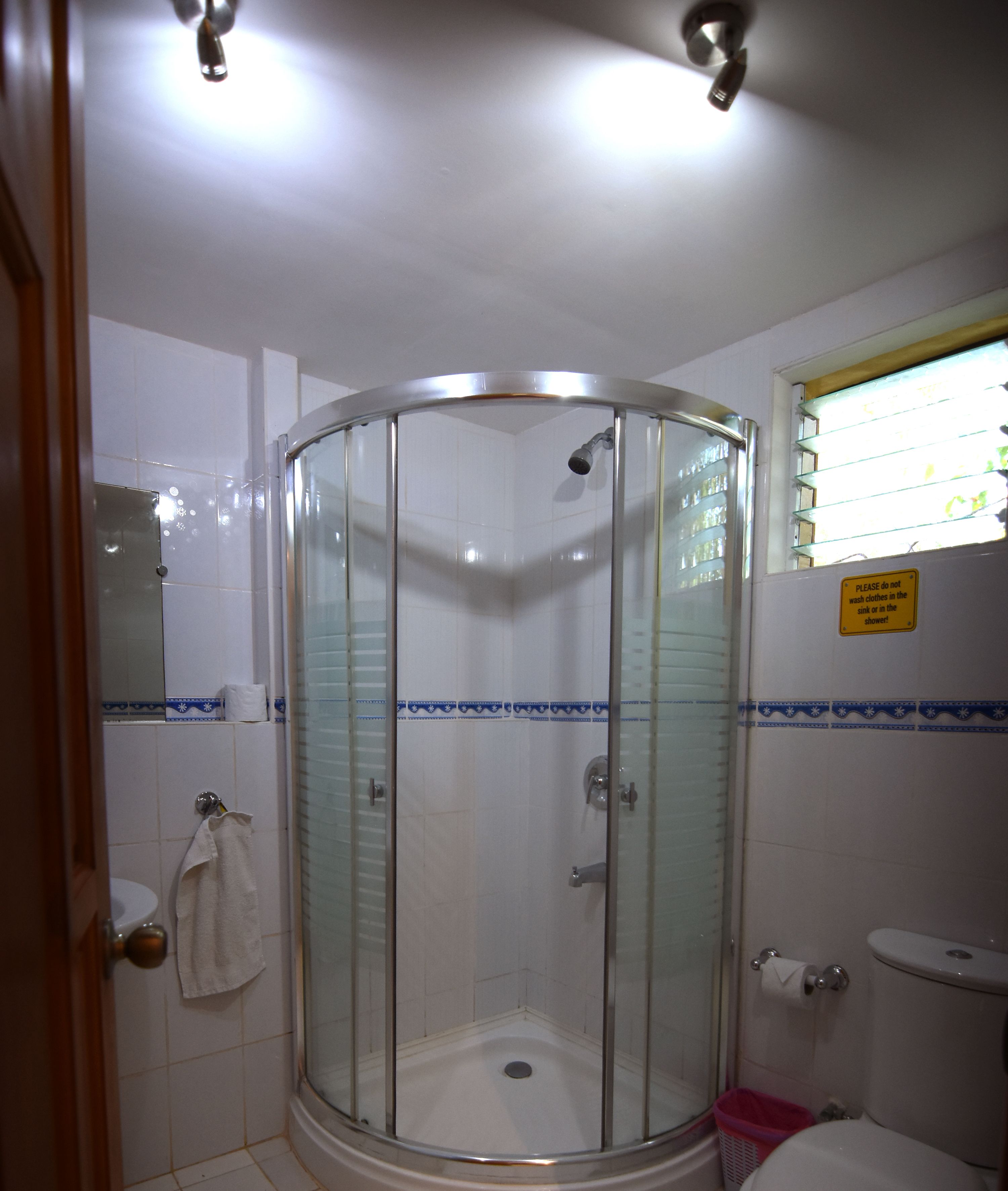 A glass-enclosed shower stall with a white tiled floor, a toilet with a pink trash bin, and a towel hanging on the door.