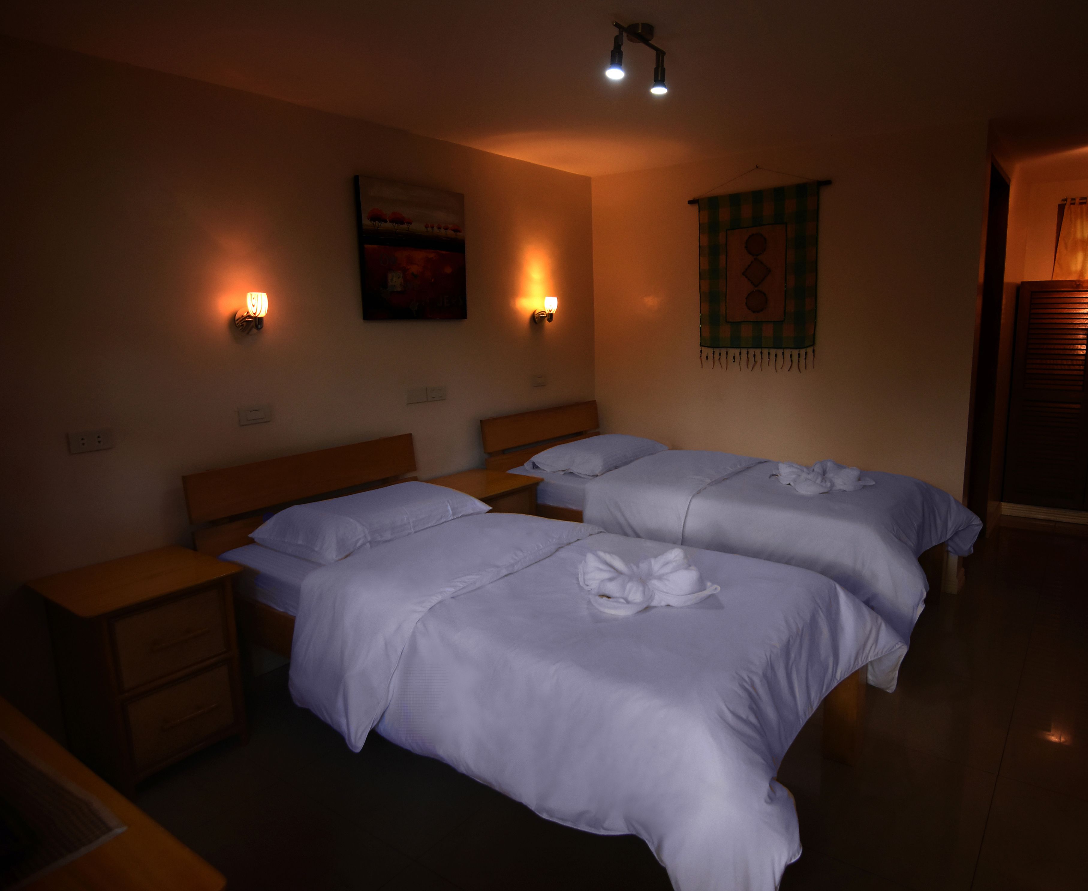A hotel room with two beds, wooden headboards, bedside tables, and wall-mounted lights.