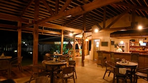 An indoor dining area with wooden tables and chairs, a bar area with a counter and shelves, and large windows allowing natural light.