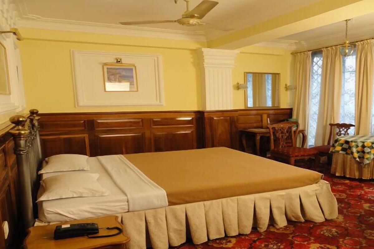 Ein Hotelzimmer mit einem großen Bett, einem Holzkopfstück, einem Schreibtisch mit Telefon, einem Sessel, einem kleinen Tisch mit kariertem Tischläufer und einem Kamin. An der Decke befindet sich ein Ventilator.