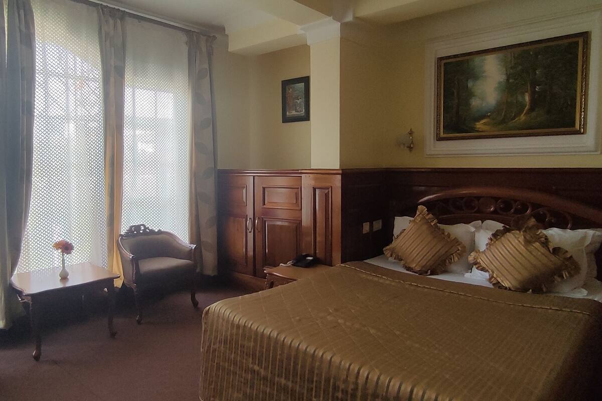 Ein Hotelzimmer mit einem großen Bett, einem Holzkopfende, einem Schreibtisch, einem Stuhl und einem Fenster mit durchsichtigen Vorhängen.