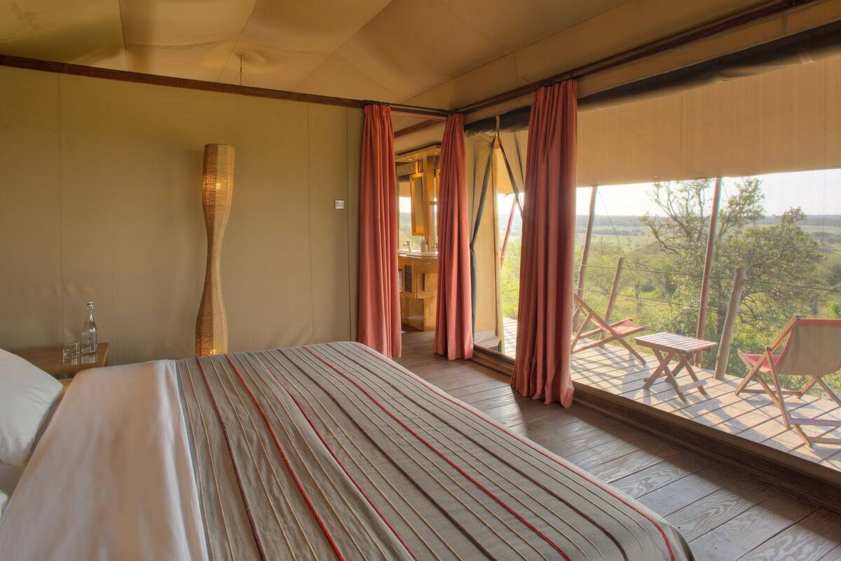 A bedroom with a large bed, a wooden floor, a view of the outdoors, and a small table with a chair.
