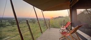 A wooden deck with two red lounge chairs and a table, overlooking a grassy landscape with a sunset in the background. - Basecamp Eagle View (Maasai Mara)