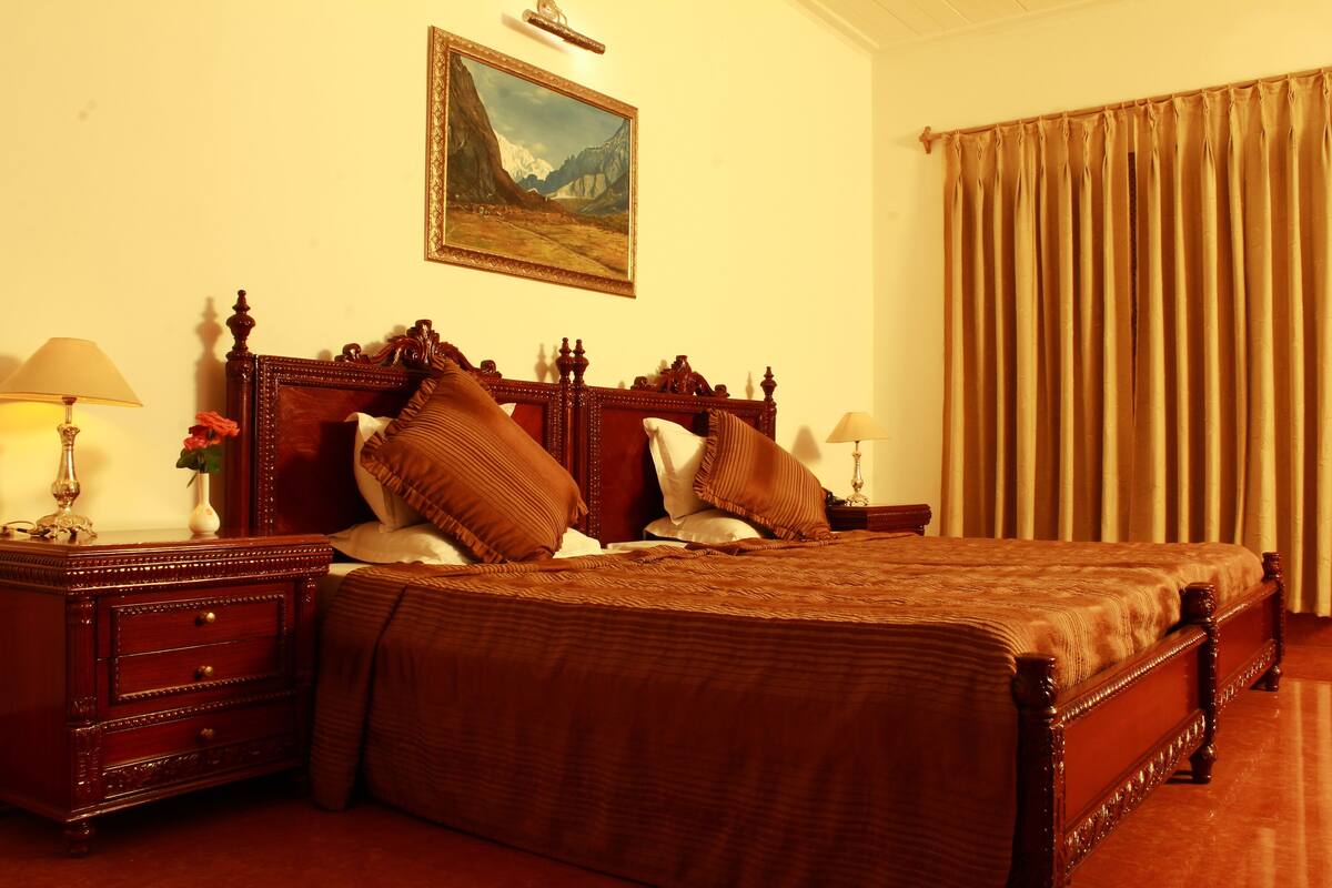 Habitación de hotel con una cama grande, cabecero de madera, dos mesitas de noche con lámparas, un cuadro en la pared y una ventana con cortinas.