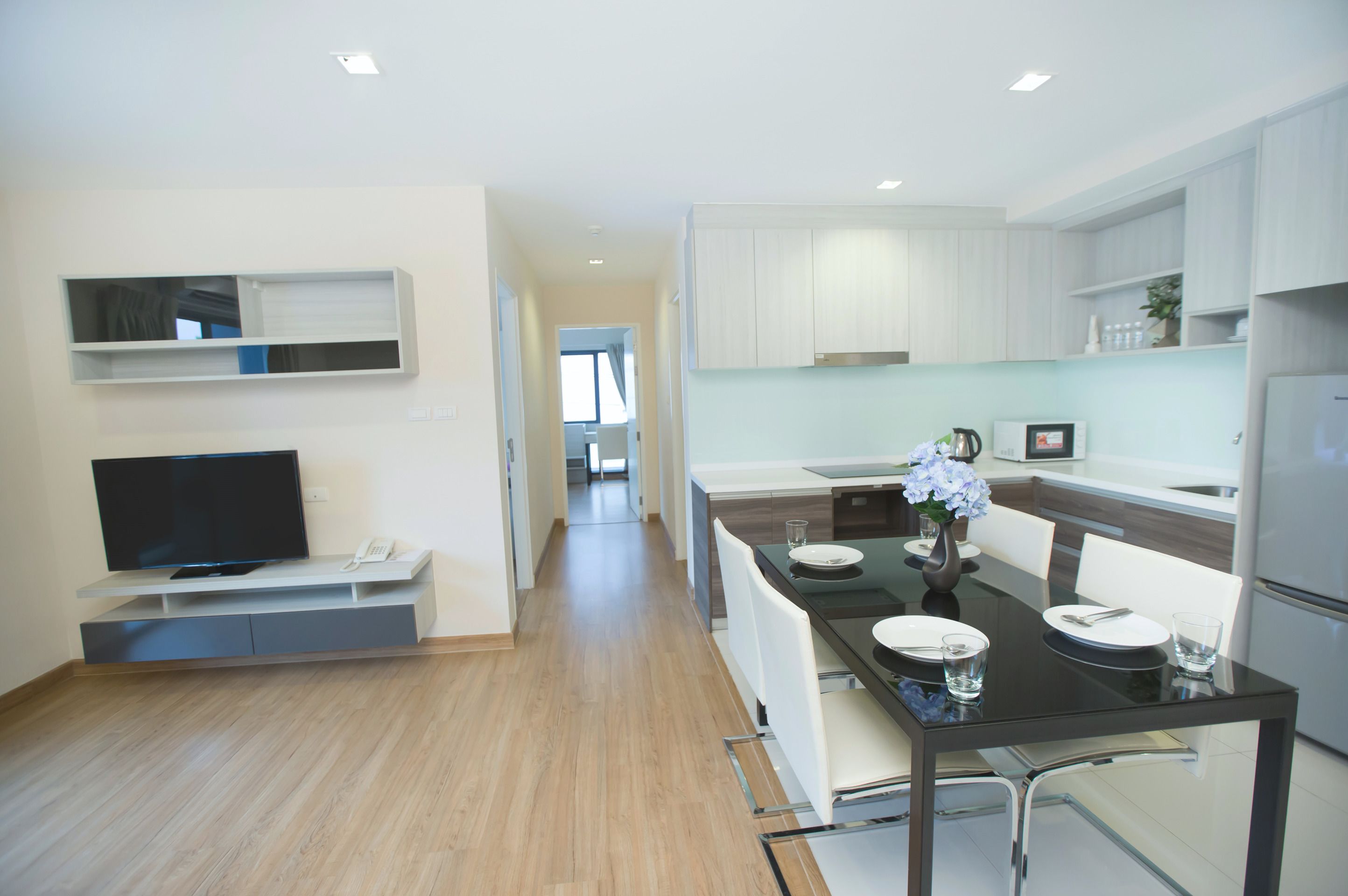 A modern kitchen with white cabinets, a dining table set for two, and a flat-screen TV on a built-in shelf.