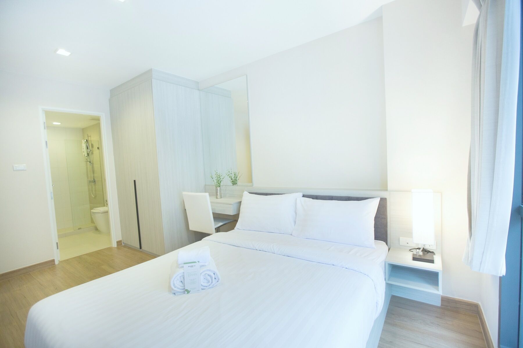 A hotel room with a large bed, a bedside table with a lamp, a bathroom with a shower and toilet, and a large mirror.