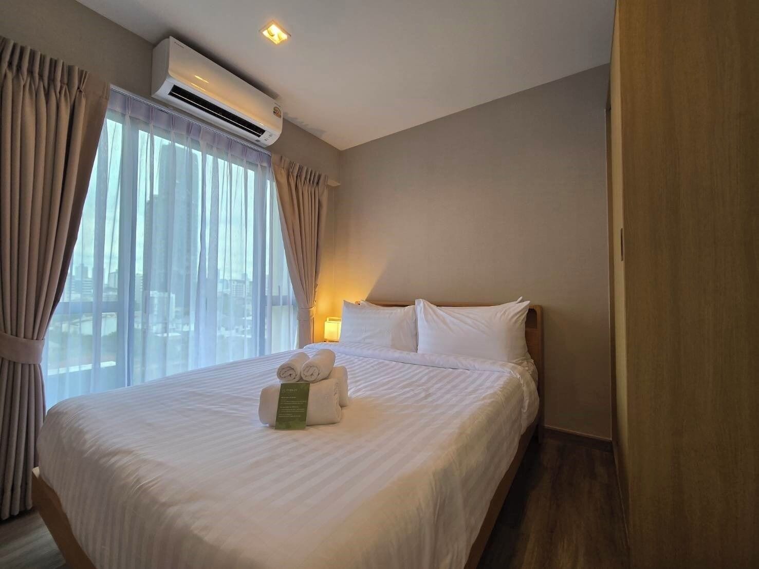 A hotel room with a large bed, an air conditioning unit, a nightstand, and a view of the city through large windows.