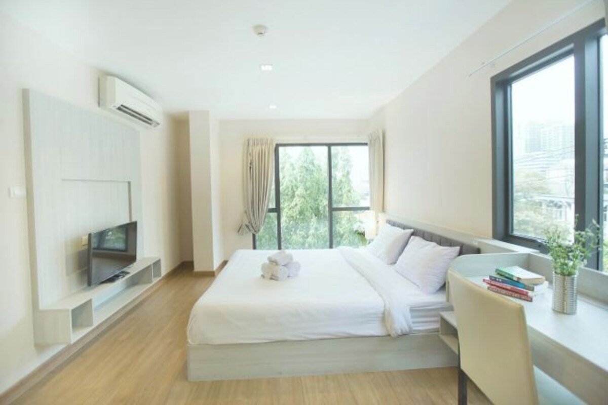 Una habitación de hotel moderna con una cama, un televisor sobre un soporte, una ventana con vistas a la vegetación y una mesita pequeña con libros y una planta.