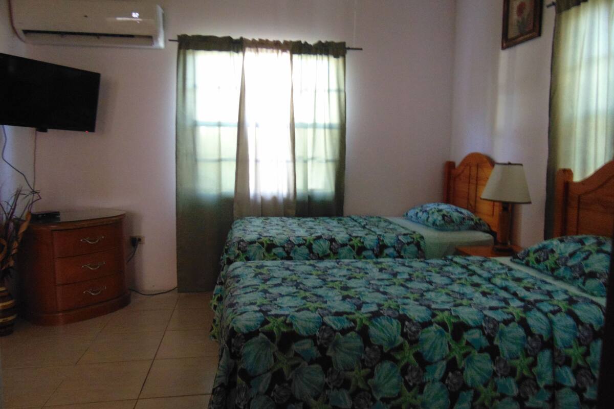 A bedroom with two beds, a dresser, a TV mounted on the wall, and a window with curtains.